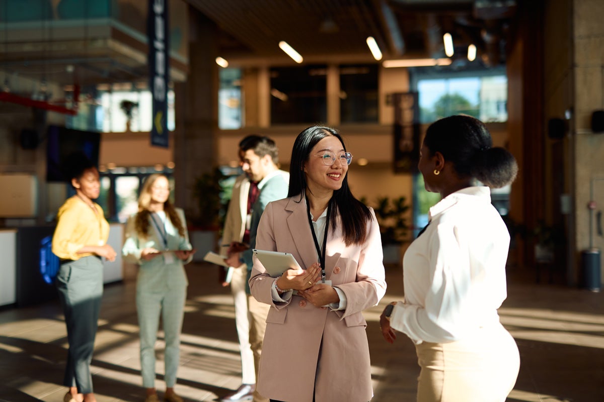 Business professionals talking and smiling in a sunlit modern office lobby, holding tablets and notebooks.