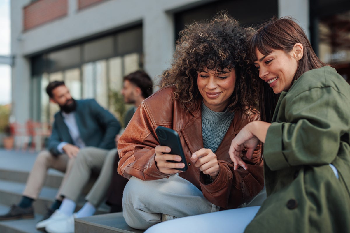 Two women smiling and looking at a phone together while two men talk in the background outdoors.