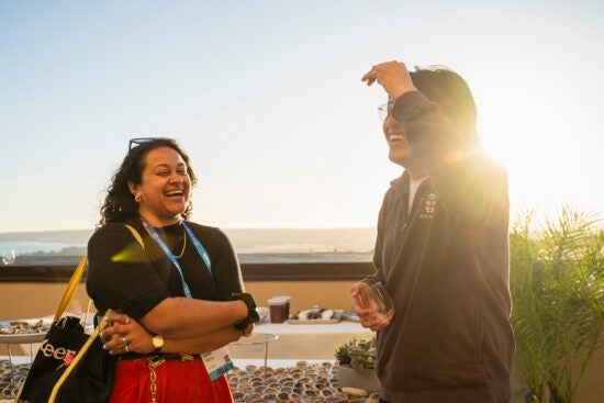 Two people laugh and talk outdoors at sunset with drinks in hand and a scenic background behind them.