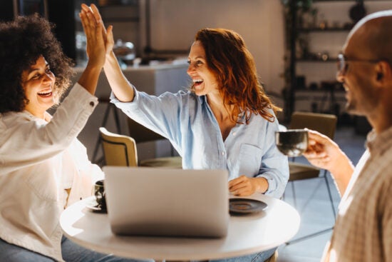 Three people at a cafe table smiling, two giving a high five, with a laptop and coffee cups in front of them.