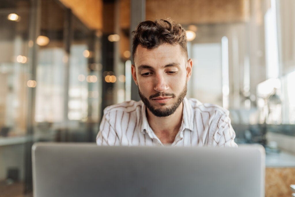 Man with a beard and striped shirt working on a laptop in a modern office with glass walls.