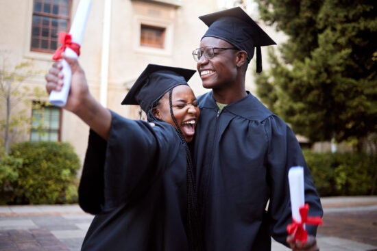 Two graduates in caps and gowns smiling and celebrating while holding their diplomas outdoors.