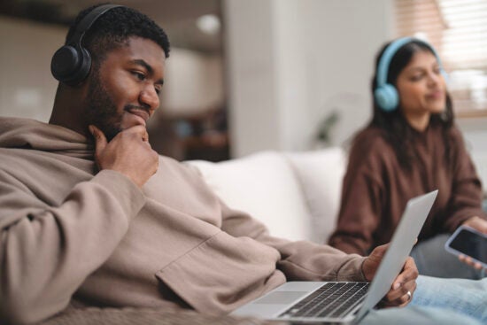 Man wearing headphones using a laptop on a couch, with a woman in headphones sitting in the background.