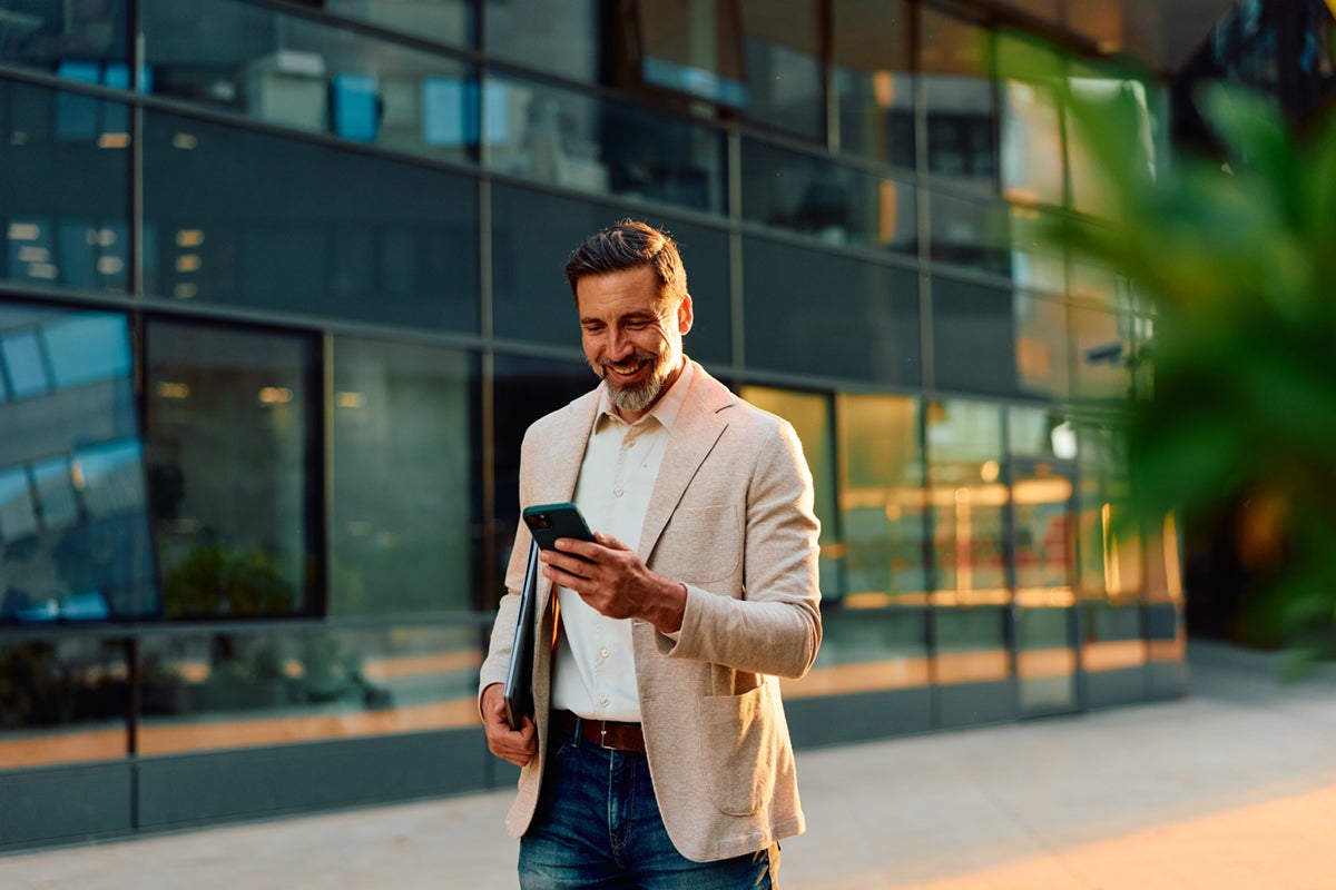 Smiling man in a light blazer walks outside a modern building while looking at his phone.