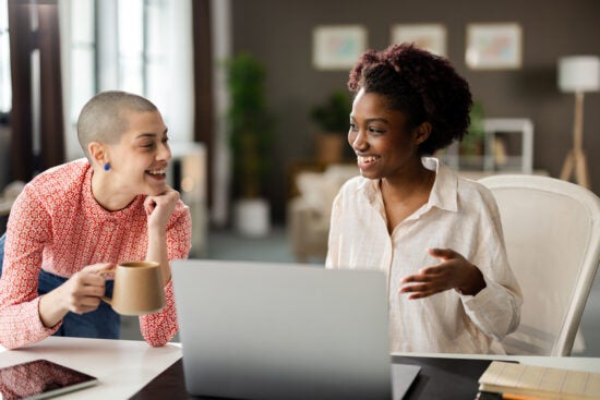 Two women smiling and talking while working together at a desk with a laptop and coffee mug.