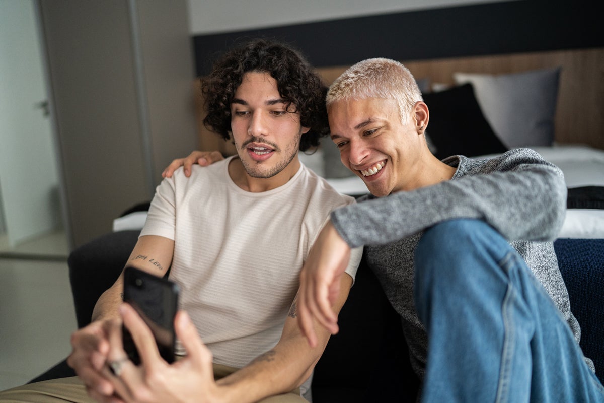Two young men sitting together on a couch, smiling and looking at a smartphone.
