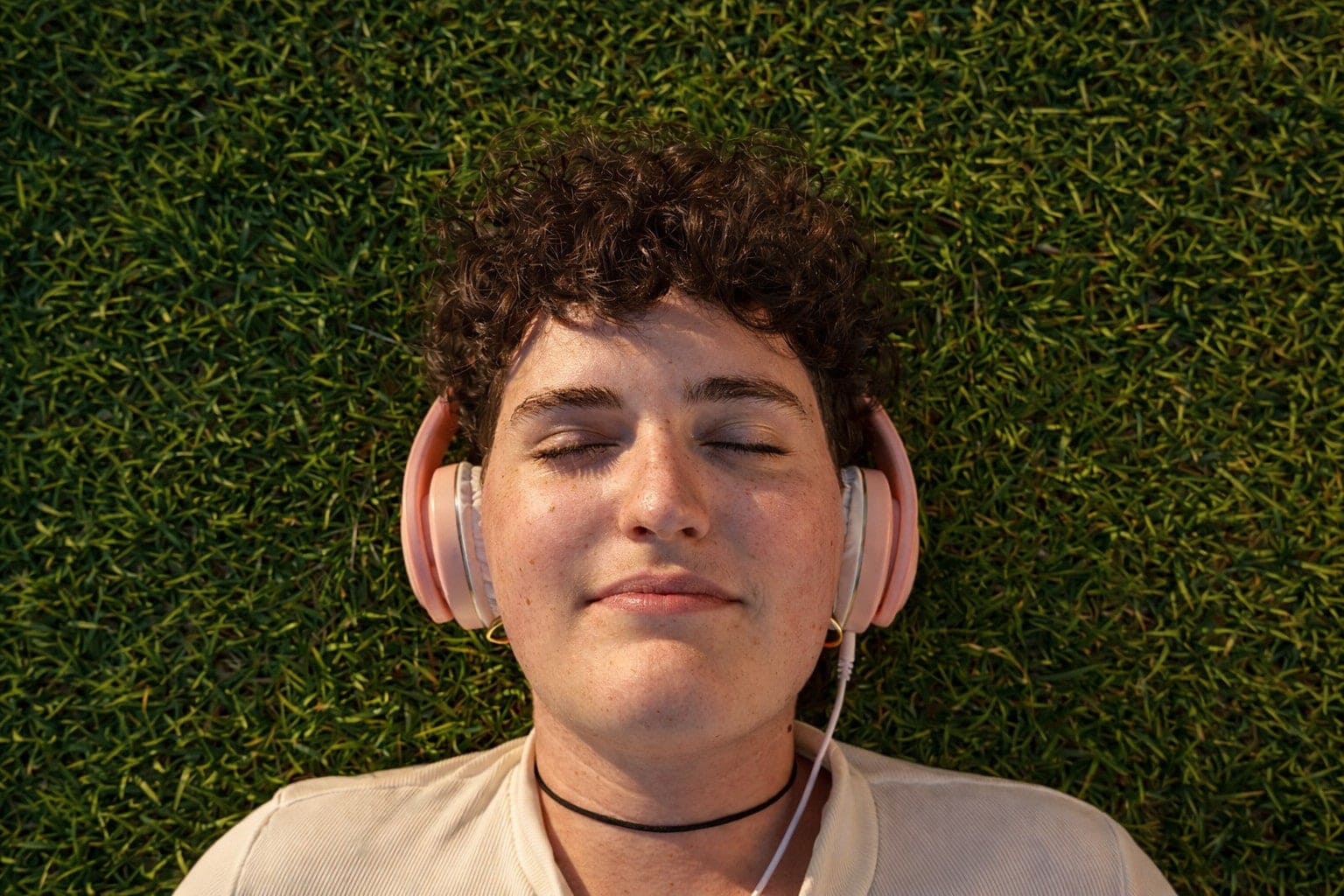 Person with curly hair lying on grass, eyes closed, wearing pink headphones and a light shirt, enjoying music.