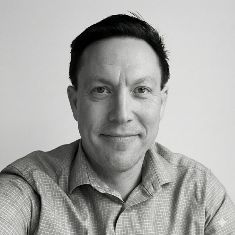Man smiling at the camera, wearing a checked shirt, in a black-and-white portrait against a plain background.
