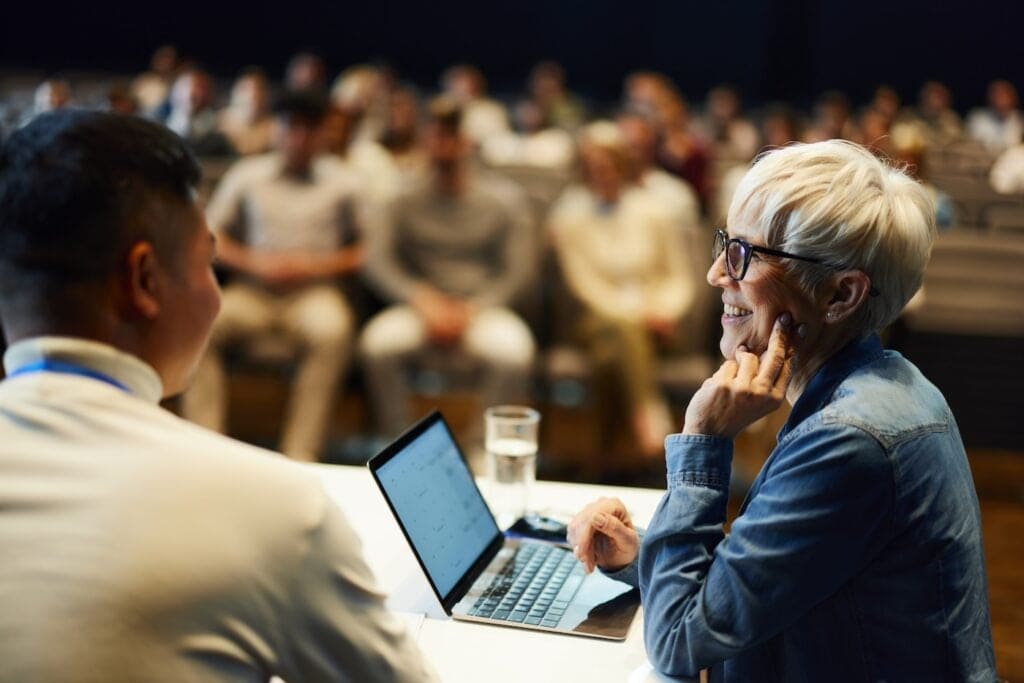 Happy senior businesswoman having panel discussion with her colleague in front of a crowd at convention center.