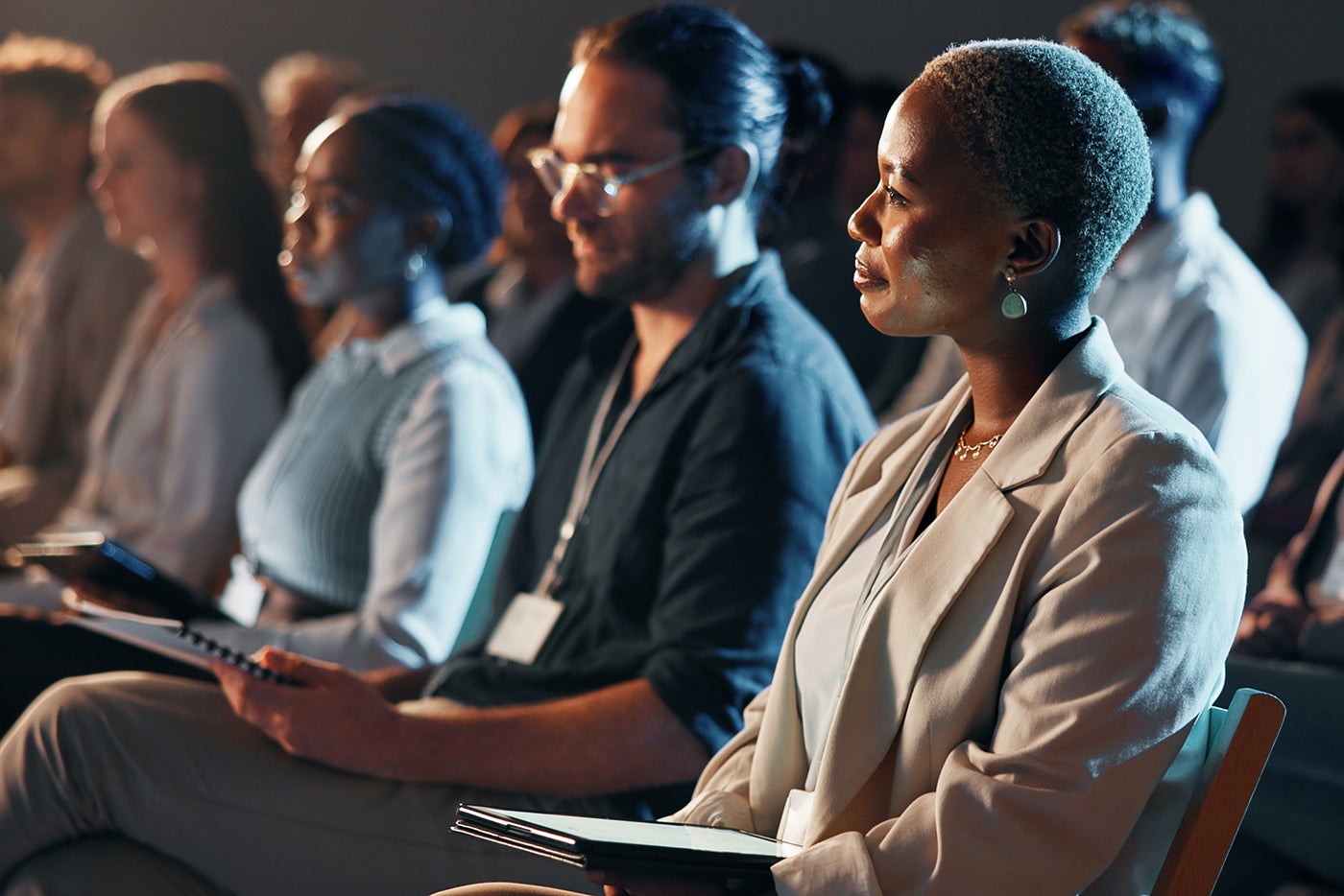 People sitting in rows at a conference, attentively listening and holding notebooks and pens.