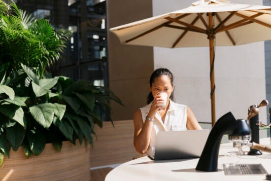 Woman sitting at an outdoor table with a laptop, drinking from a paper cup under a large umbrella.