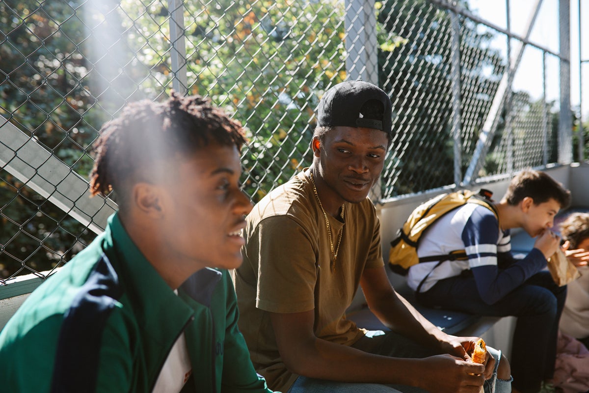 Two young men sit on bleachers outdoors, talking, with others in the background near a chain-link fence.