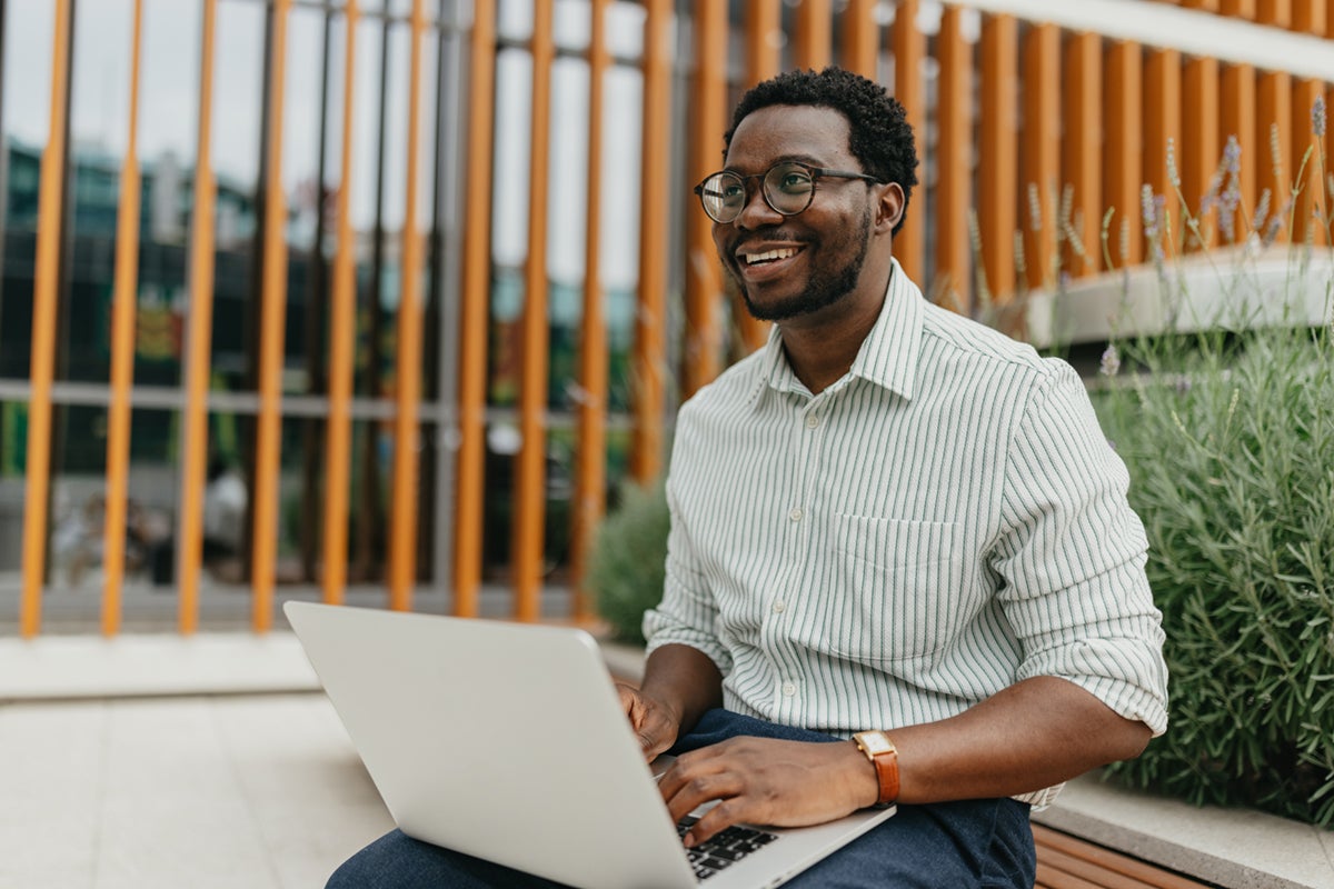 Smiling man with glasses works on a laptop while sitting outside near plants and modern wooden panels.