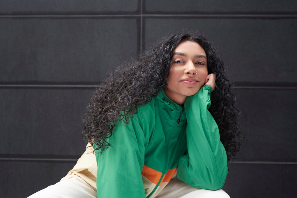 Woman with long curly hair wearing a green jacket, resting her chin on her hand and smiling slightly.