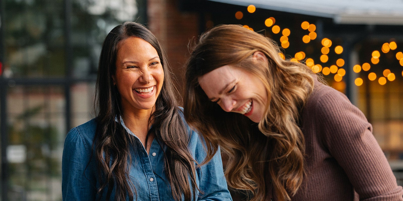 Two women laughing together outdoors under branded string lights, with the warm glow blurred in the background.