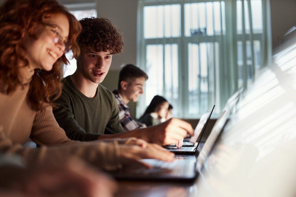 Four students sit side by side at desks in a bright classroom, working on laptops. Two appear to be collaborating and smiling, while others focus on their screens.