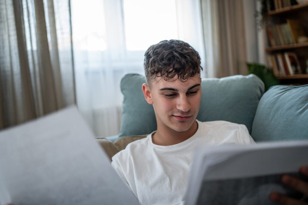 A young man sits on a couch, smiling while reading papers. Light comes through the window behind him, and bookshelves are visible in the background.