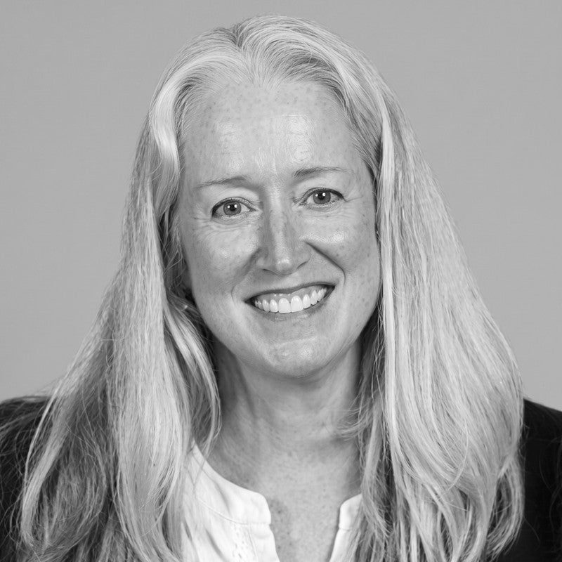 A woman with long, light hair smiles at the camera. She is wearing a light blouse and dark jacket. The photo is black and white with a plain background.