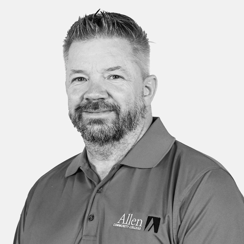 A man with short hair and a beard, wearing a polo shirt with the “Allen Community College” logo, smiles at the camera against a plain background.