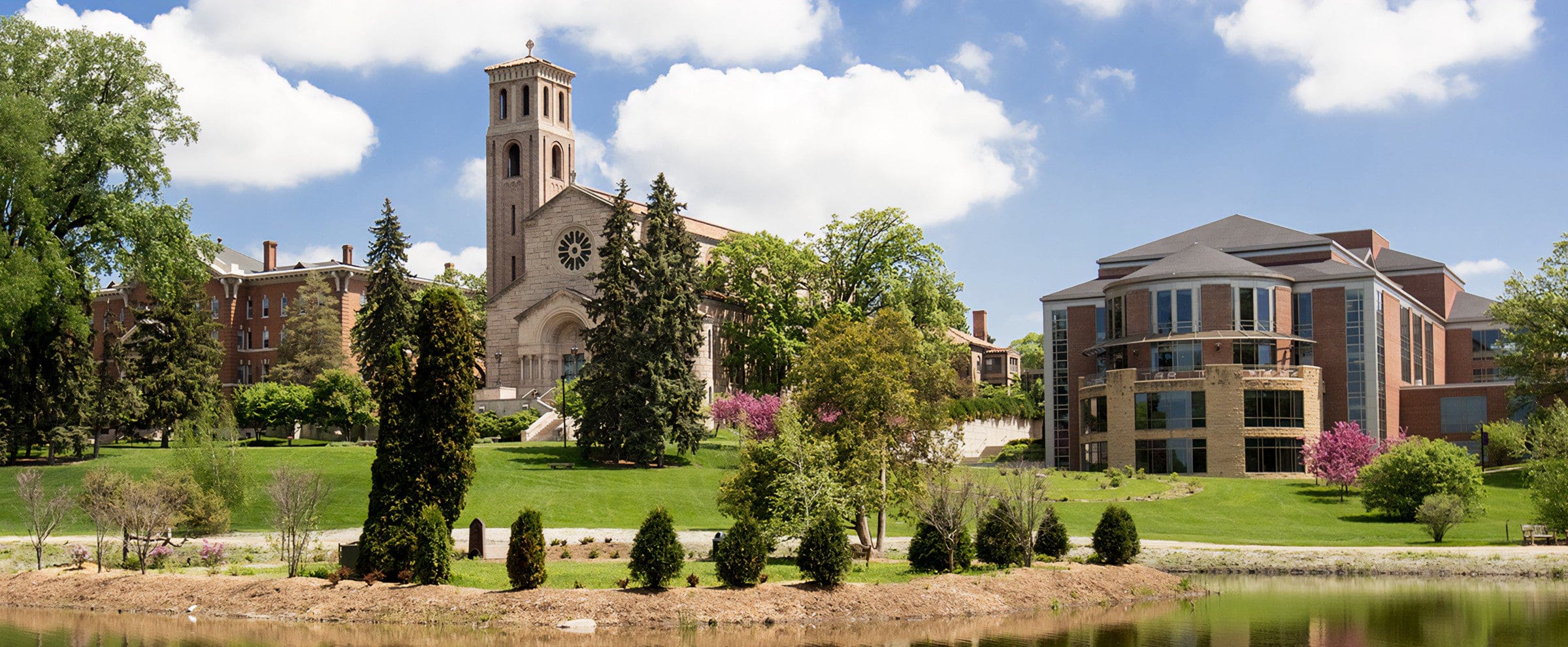 A campus scene with a historic church, modern building, trees, and a pond under a partly cloudy sky.