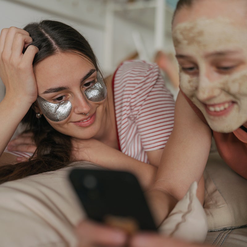 Two people relax indoors, one with silver under-eye patches and the other with a face mask. They are lying down, smiling, and looking at a smartphone together.
