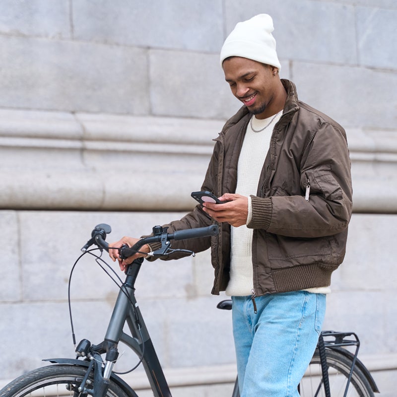 A man in a brown jacket and white beanie stands beside a bicycle, smiling while looking at his phone. Stone wall in the background.