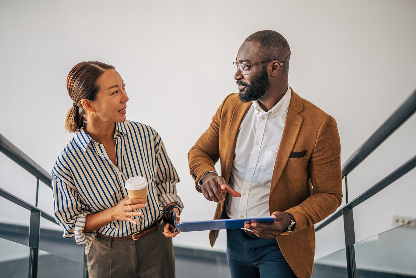 Two professionals talking on stairs; one holds a coffee cup, the other holds a clipboard and gestures while they converse in a modern office setting.