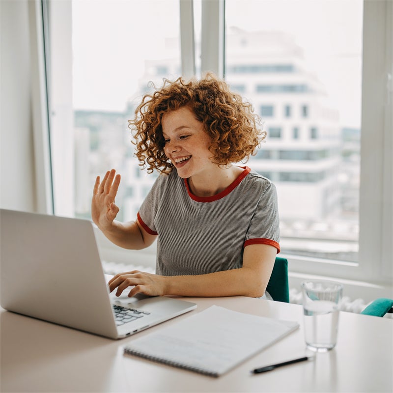 A smiling person with curly hair waves at a laptop during a video call, sitting at a desk with a notebook, pen, and glass of water by a window with city buildings outside.
