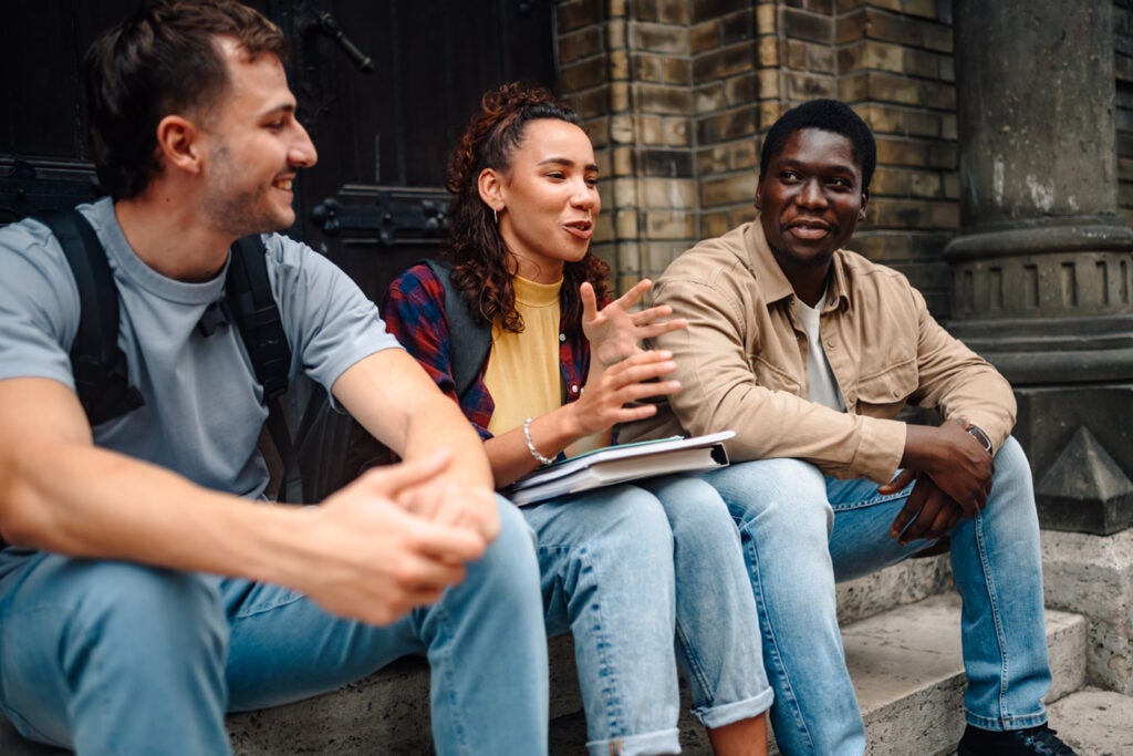 Three young adults sit on stone steps outside, chatting and smiling. One person gestures while holding a notebook. They all appear relaxed and engaged in conversation.