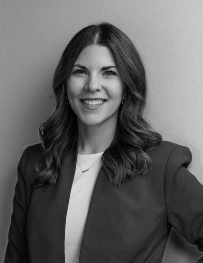 Black and white portrait of a woman with wavy hair, wearing a blazer over a light top, smiling at the camera against a plain background.