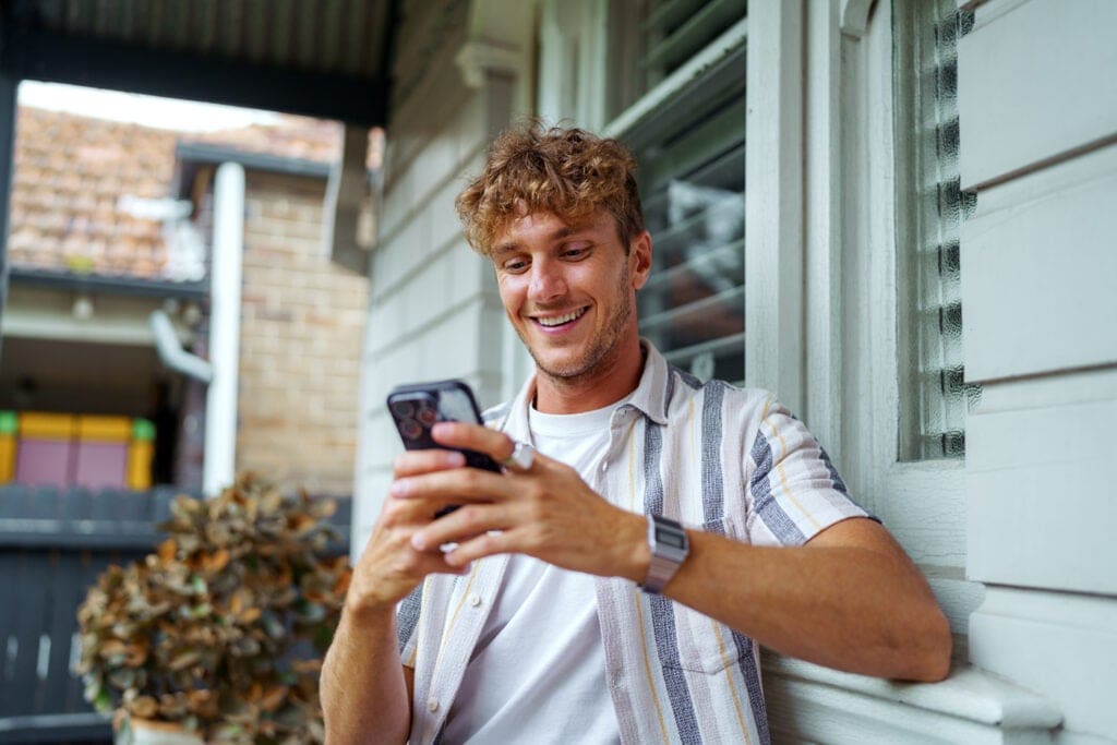 Young man with curly hair smiles while looking at his smartphone, sitting outside by a house window.