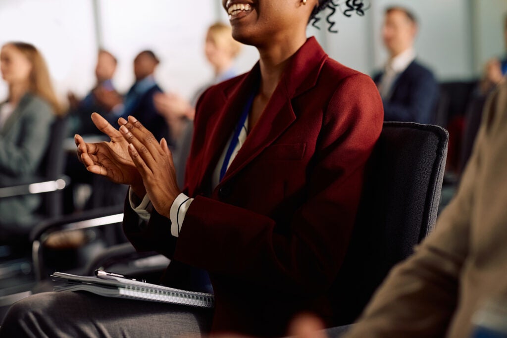 A woman in a burgundy blazer clapping in a conference room with other attendees in the background.