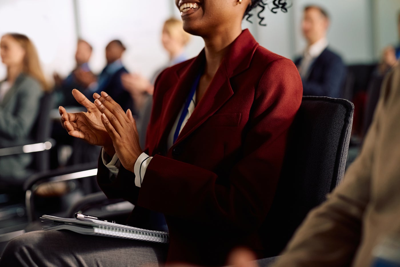 A woman in a burgundy blazer clapping in a conference room with other attendees in the background.